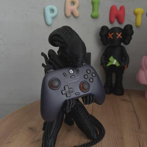 May include: A black Xenomorph figurine standing in a crouched position, holding a dark gray GameSir video game controller. The controller features orange accents on the analog sticks, a directional pad, and various buttons. The figure has a long tail.