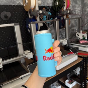May include: A light blue can cooler with the Red Bull logo in red and yellow. The cooler has a handle and is designed to hold a standard-sized beverage can. The background shows 3D printers and spools of filament.