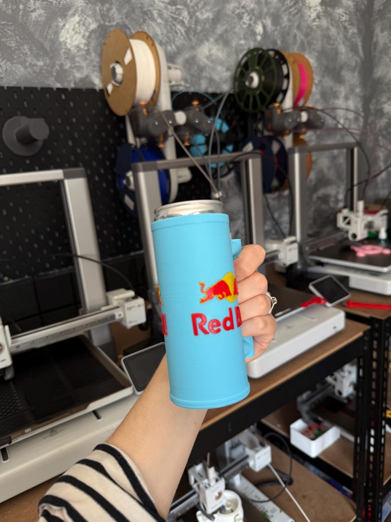 May include: A light blue can cooler with the Red Bull logo in red and yellow. The cooler has a handle and is designed to hold a standard-sized beverage can. The background shows 3D printers and spools of filament.