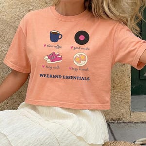 Może przedstawiać: Brzoskwiniowy t-shirt z krótkim rękawem z nadrukiem graficznym przedstawiającym cztery ikony: niebieski kubek do kawy, czarną płytę winylową, różowe trampki i dwa jajka sadzone. Tekst głosi: slow coffee, good music, long walk, lazy brunch, WEEKEND ESSENTIALS.