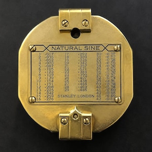 Puede incluir: Un instrumento circular de latón con una placa rectangular central grabada con el texto NATURAL SINE y STANLEY LONDON, junto a una tabla de valores numéricos. La carcasa de latón incluye dos soportes de montaje asegurados con tornillos.
