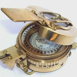 May include: A vintage brass compass with a hinged lid and a sighting mechanism. The compass face features degree markings and directional indicators. The open lid reveals a magnifying lens. The compass is designed for navigation and direction finding.