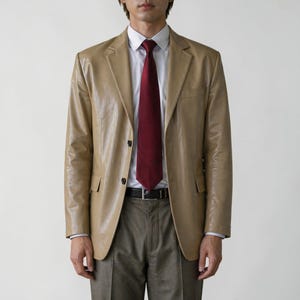 May include: A tan leather blazer with a burgundy tie, white dress shirt, and gray trousers. The blazer has two black buttons and two front pockets. The outfit is completed with a black belt.