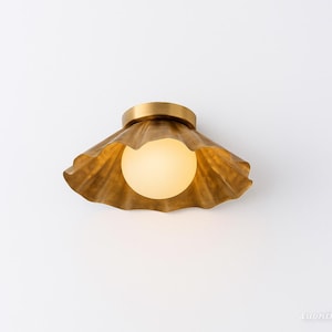 May include: A gold-colored, seashell-shaped ceiling light fixture with a round, frosted glass bulb. The fixture is mounted on a white wall. The light has a decorative, wavy design.