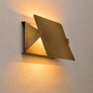 May include: A wall-mounted light fixture made of brushed brass with a geometric, angular design. The fixture features a flat, rectangular front plate angled outward, emitting a warm, soft glow from behind the metal panels onto the wall.