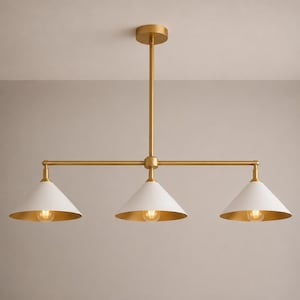 May include: A linear chandelier featuring a central brass rod and three conical shades. Each shade has a matte white exterior and a metallic gold interior, housing a single light bulb. The fixture is suspended by a matching brass ceiling mount.