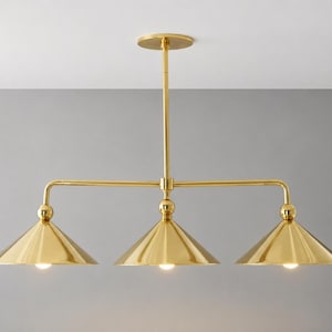 Peut inclure: Une lampe suspendue à trois lumières de couleur or avec des abat-jours en forme de cône. Le luminaire est composé d'une tige verticale centrale fixée à une barre horizontale avec trois lumières orientées vers le bas. Les abat-jours sont dorés brillants.