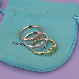 May include: Three open-ended rings in rose gold, silver, and yellow gold tones, each featuring a T-shaped design at the opening. The rings are resting on a light blue fabric pouch with a drawstring closure and the text Tiffany & Co. printed in black.