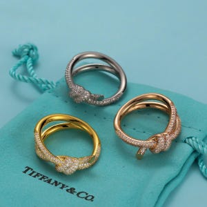 May include: Three rings featuring a knot design and pavé-set stones. One ring is silver-toned, one is gold-toned, and one is rose gold-toned. The rings are displayed on a light blue fabric pouch with the text Tiffany & Co. printed in black.
