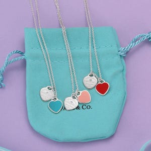 May include: Three silver necklaces with heart-shaped pendants, each featuring a small round diamond and a coloured enamel heart charm in turquoise, light pink, or red. The pendants are engraved with Tiffany & Co. and rest on a turquoise drawstring pouch.