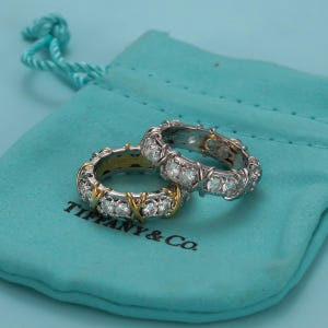 May include: Two rings featuring a silver-tone band with a repeating X-shaped design in gold-tone metal, accented with clear round stones. The rings are resting on a light blue fabric pouch with the text TIFFANY & CO. printed in black ink.