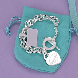 Op de afbeelding: Een schakelarmband van sterling zilver met een knevelsluiting en een hartvormig label met de inscriptie Please Return To Tiffany & Co. New York 925. De armband ligt op een klein, lichtblauw stoffen zakje met een trekkoord.