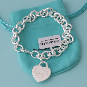 Op de afbeelding: Een schakelarmband van sterling zilver met een hartvormige hanger gegraveerd met de tekst PLEASE RETURN TO TIFFANY & CO. NEW YORK 925. De armband bevat een wit rechthoekig label met de tekst TIFFANY & CO. NEW YORK SINCE 1837.