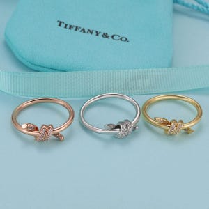 May include: Three rings in rose gold, silver, and yellow gold tones, each featuring a knot design embellished with small clear stones. The rings are positioned in front of a light blue fabric pouch with the text Tiffany & Co. printed in black.