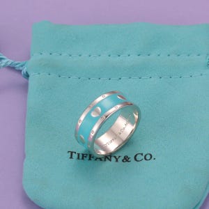 May include: A sterling silver band ring featuring a central inlay of light blue enamel with repeating heart motifs and small circular accents. The interior of the band is engraved with the text TIFFANY & CO. The ring rests on a light blue fabric pouch.