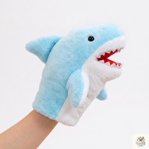 May include: A plush shark hand puppet featuring light blue fur on the top and white fur on the underside. The shark has a wide open mouth with a red interior and white felt teeth, along with a small dorsal fin and black plastic eyes.
