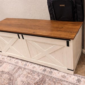 May include: A wooden storage chest with a light brown top and white body. The chest features a farmhouse style with black metal accents and an X-shaped design on the front. A black bag rests on top.