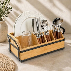May include: A wooden cutlery and dish drying rack with black metal accents. The rack holds plates, a glass, and silverware with wooden handles. A vase with greenery is in the background.