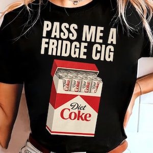 May include: Black t-shirt with the text "PASS ME A FRIDGE CIG" in white block letters. Below the text is a graphic of a Diet Coke carton with six Diet Coke cans inside. The Diet Coke logo is visible on the carton.