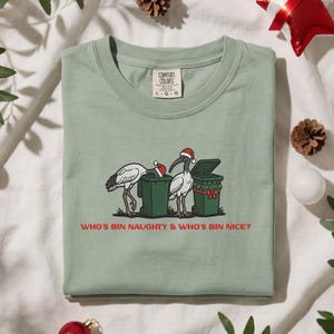 May include: A sage green t-shirt featuring a graphic of two white birds wearing Santa hats, each standing by a green trash bin. One bin is decorated with a red bow and holly. The text reads: WHO'S BIN NAUGHTY & WHO'S BIN NICE? Size L.