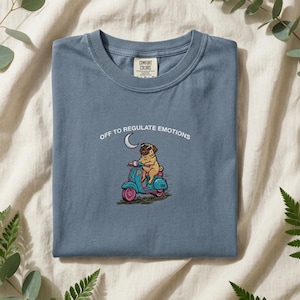 Può includere: T-shirt blu con la scritta "OFF TO REGULATE EMOTIONS" e un disegno di un carlino su uno scooter turchese. Il carlino indossa occhiali da sole e un cuore fluttua sopra lo scooter. La maglietta ha un'etichetta Comfort Colors.