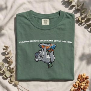 May include: A sage green t-shirt featuring a graphic of a grey koala wearing a beanie and climbing a rock wall. The text above the koala reads, "CLIMBING BECAUSE DRUGS CAN'T GET ME THIS HIGH." The shirt has a crew neckline and a Comfort Colors brand label.
