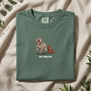 May include: Green crew neck t-shirt with a monkey graphic. The design shows two monkeys, one standing and one sitting. The words "BE BRAVE" are printed below. The shirt also has the "COMFORT COLORS" logo.