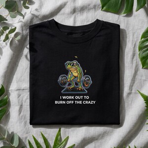 Könnte beinhalten: Schwarzes T-Shirt mit einem Cartoon-Frosch, der eine Sonnenbrille trägt und Gewichte hebt. Der Frosch ist grün und gelb, mit dem Text "I WORK OUT TO BURN OFF THE CRAZY" darunter. Das T-Shirt liegt gefaltet auf einer hellgrauen Oberfläche.