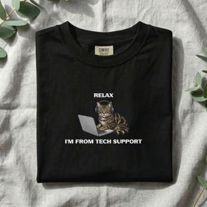 May include: A black crew neck t-shirt featuring a graphic of a brown tabby cat wearing a headset while sitting at a laptop. The text above the cat reads RELAX and the text below the cat reads I'M FROM TECH SUPPORT. The shirt has a Comfort Colors label.