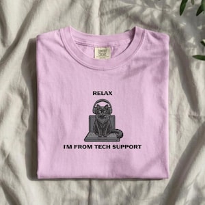 May include: A light purple crew neck t-shirt featuring a black graphic of a cat wearing headphones while sitting at a laptop. The text above the cat reads RELAX and the text below reads I'M FROM TECH SUPPORT. The shirt has a Comfort Colors label.