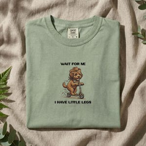 Op de afbeelding: Een saliegroen t-shirt met een afbeelding van een bruine hond met een cowboyhoed en bandana op een step. De tekst boven de hond luidt WAIT FOR ME en de tekst eronder luidt I HAVE LITTLE LEGS. Het shirt is voorzien van een Comfort Colors label.