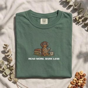 Puede incluir: Una camiseta verde salvia con un gráfico de un perro salchicha marrón con gafas redondas, leyendo un libro abierto junto a una pila de libros y una taza de café. El texto debajo del gráfico dice: READ MORE, BARK LESS.