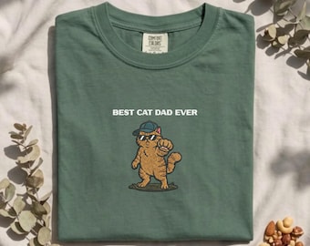 Puede incluir: Una camiseta de color verde salvia con el gráfico de un gato atigrado naranja que lleva una gorra azul y gafas de sol negras, con el texto "BEST CAT DAD EVER" impreso en blanco sobre la ilustración del gato. La camiseta tiene cuello redondo.