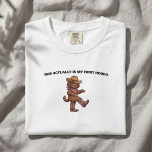 May include: A white t-shirt with a graphic of a brown dog wearing a cowboy hat, a pink bandana, and pink cowboy boots. The text above the dog reads, THIS ACTUALLY IS MY FIRST RODEO. The shirt has a Comfort Colors brand label at the inner neckline.
