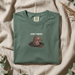 Könnte beinhalten: Ein salbeigrünes T-Shirt mit Rundhalsausschnitt und einer Grafik eines braunen Maulwurfs, der eine kleine Schaufel hält und aus einem Loch im Boden kommt. Über dem Maulwurf ist der Text DIRT NERD in Weiß gedruckt. Das Shirt hat ein Comfort Colors Etikett.