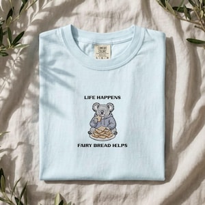 May include: A light blue t-shirt featuring a graphic of a grey koala wearing a hoodie, eating a piece of toast, with a stack of toast on a plate. The text above the koala reads LIFE HAPPENS and the text below reads FAIRY BREAD HELPS.