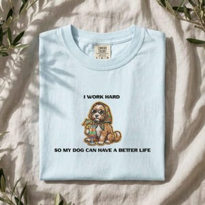 Peut inclure: Un t-shirt bleu clair avec une illustration d'un chien brun portant un foulard et des lunettes de soleil, tenant une boisson tropicale. Le texte au-dessus du chien indique I WORK HARD et le texte en dessous indique SO MY DOG CAN HAVE A BETTER LIFE.