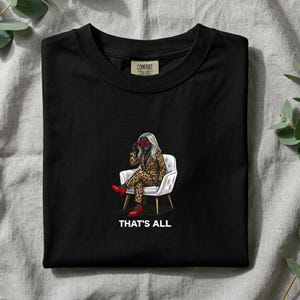 Puede incluir: Una camiseta negra con un gráfico de una persona de pelo blanco con un traje de estampado de leopardo y gafas rojas, sentada en una silla blanca. La persona lleva tacones rojos. El texto "THAT'S ALL" está impreso en blanco debajo del gráfico.