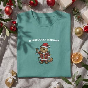 May include: A teal crew neck t-shirt featuring a graphic of a grumpy brown cat wearing a Santa hat and a red suit, tangled in colourful Christmas lights. Above the cat, the white text reads: IS THIS JOLLY ENOUGH? The shirt has short sleeves.