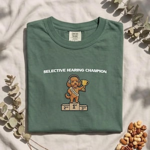 May include: A sage green t-shirt featuring a graphic of a brown dog wearing headphones, standing on a first-place podium while holding a gold trophy. The text above the graphic reads SELECTIVE HEARING CHAMPION in white capital letters.
