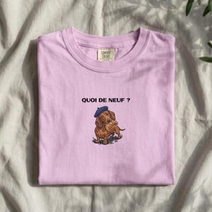 May include: A light purple t-shirt featuring a graphic of a brown dachshund wearing a blue beret and holding a croissant in its mouth. Above the dog, the text reads "QUOI DE NEUF ?" in black letters. The shirt has a Comfort Colors brand label.