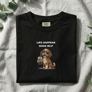 Peut inclure: Un t-shirt noir avec une illustration d'un chien marron portant des lunettes de soleil et tenant un gobelet en plastique avec une paille. Le texte au-dessus du chien indique "LIFE HAPPENS DOGS HELP" en lettres blanches. Étiquette Comfort Colors.