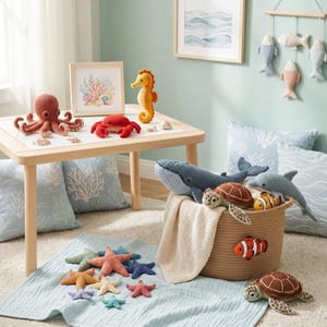 May include: A collection of handmade crochet sea creatures, including an octopus, crab, seahorse, whale, dolphins, turtles, and starfish. The toys are in various colors, such as red, orange, blue, and brown. The scene is set in a room with a light blue and white color scheme.