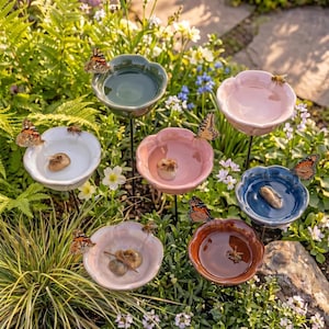 May include: A selection of flower-shaped ceramic bird baths in shades of white, pink, blue, and brown. Butterflies and bees are seen on the edges of the baths, which are set amongst greenery and flowers. The bird baths are mounted on black metal stems.