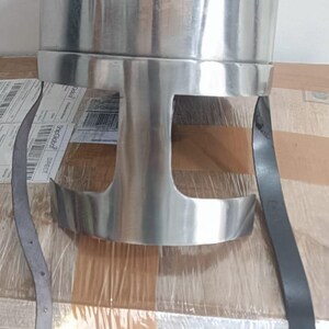 May include: A polished silver-colored metal helmet featuring a rounded dome top and a T-shaped face opening. The helmet includes two long black leather chin straps attached to the sides, with one strap featuring a metal buckle for secure fastening.