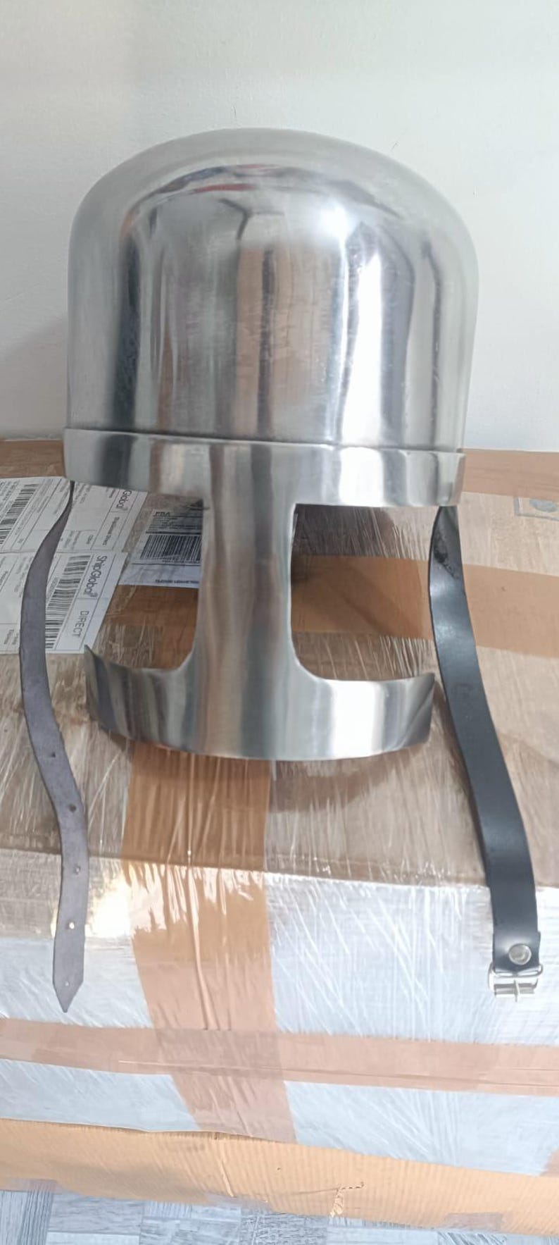 May include: A polished silver-colored metal helmet featuring a rounded dome top and a T-shaped face opening. The helmet includes two long black leather chin straps attached to the sides, with one strap featuring a metal buckle for secure fastening.