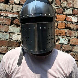 Può includere: Un elmo medievale color argento con una parte superiore nera e una visiera con piccoli fori. L'elmo ha rivetti lungo la parte superiore e un cinturino in pelle. Lo sfondo è un muro di mattoni usurato.