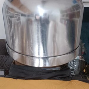 May include: A polished, reflective silver-toned metal helmet with a smooth, rounded dome shape and a distinct horizontal seam encircling the lower portion. The surface is highly reflective, showing distorted environmental light reflections.