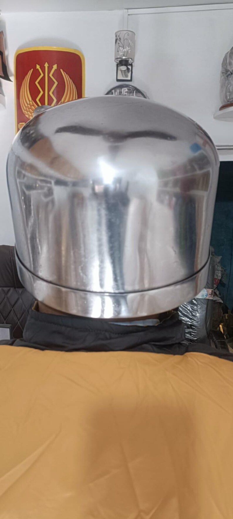 May include: A polished, reflective silver-toned metal helmet with a smooth, rounded dome shape and a distinct horizontal seam encircling the lower portion. The surface is highly reflective, showing distorted environmental light reflections.