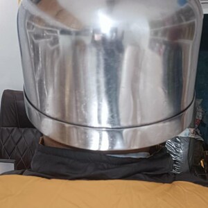 May include: A polished, silver-colored metal helmet with a smooth, rounded dome shape and a distinct horizontal seam encircling the lower portion. The surface is highly reflective, showing distorted light reflections across its curved exterior.